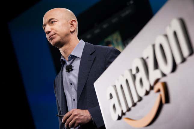 Jeff Bezos holds a phone and looks forward, an Amazon logo is on the podium next to him